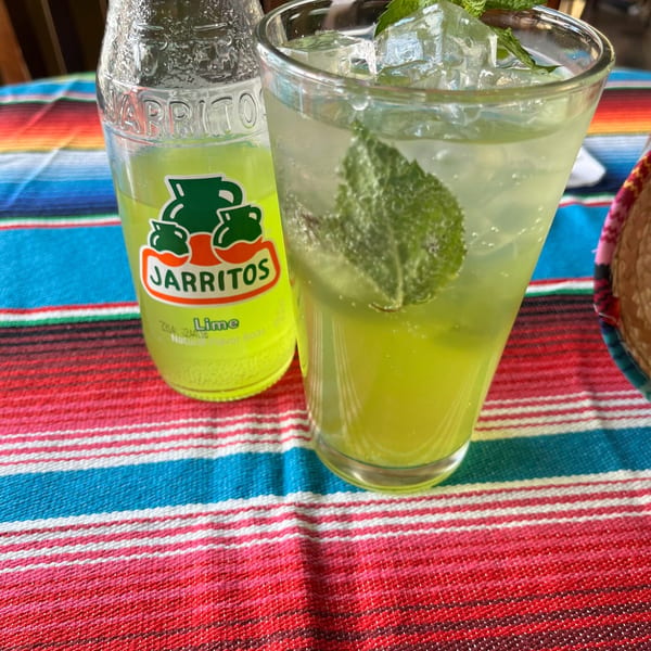 A Jarritos lime soda bottle and a glass of lime drink with mint and ice on a striped tablecloth.