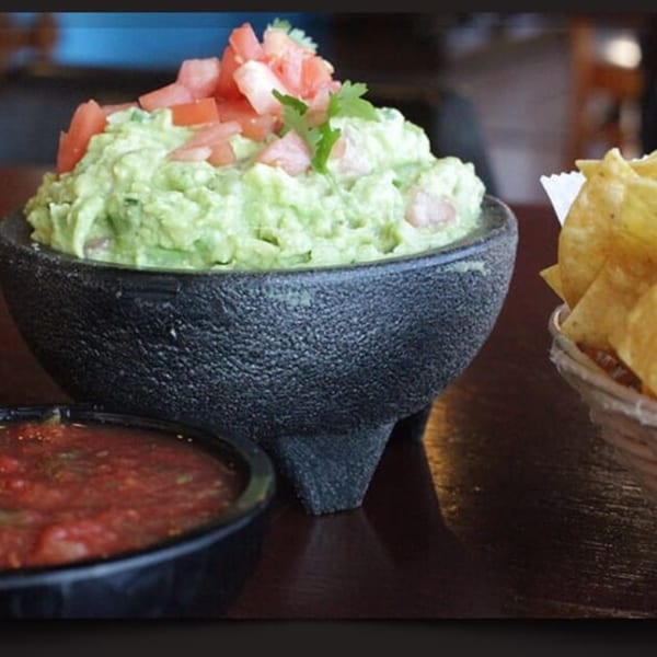 Guacamole topped with diced tomatoes and cilantro, with salsa and tortilla chips on a wooden table.
