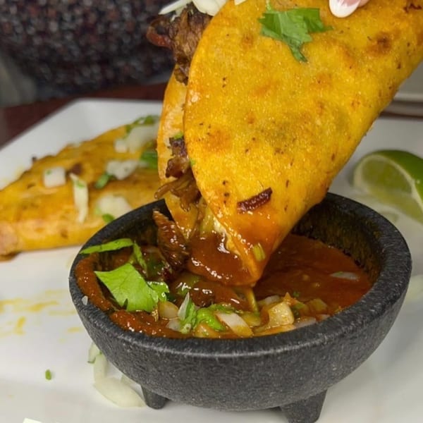 A hand dips a golden-fried birria taco into a black molcajete bowl filled with consommé.