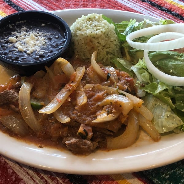 A Mexican dish with meat and onions in sauce, rice, black beans with cheese, and a side salad.