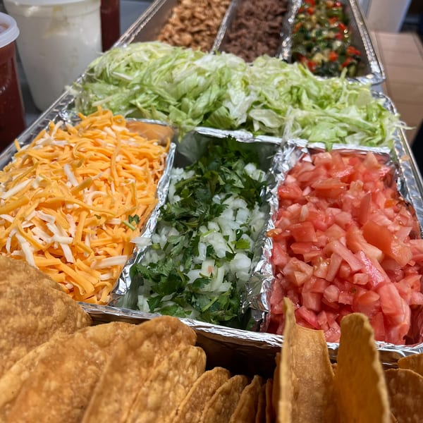 A taco bar with shredded cheese, lettuce, diced tomatoes, onions, cilantro, and crispy taco shells.