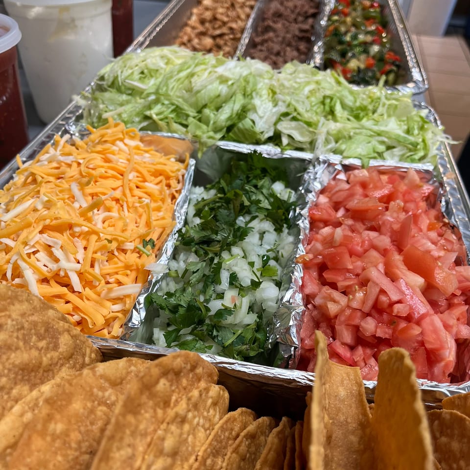 A taco bar with shredded cheese, lettuce, diced tomatoes, onions, cilantro, and crispy taco shells.