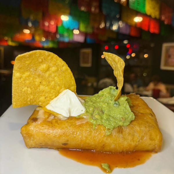 A golden-brown chimichanga topped with guacamole, sour cream, and tortilla chips, with colorful decorations in the background.