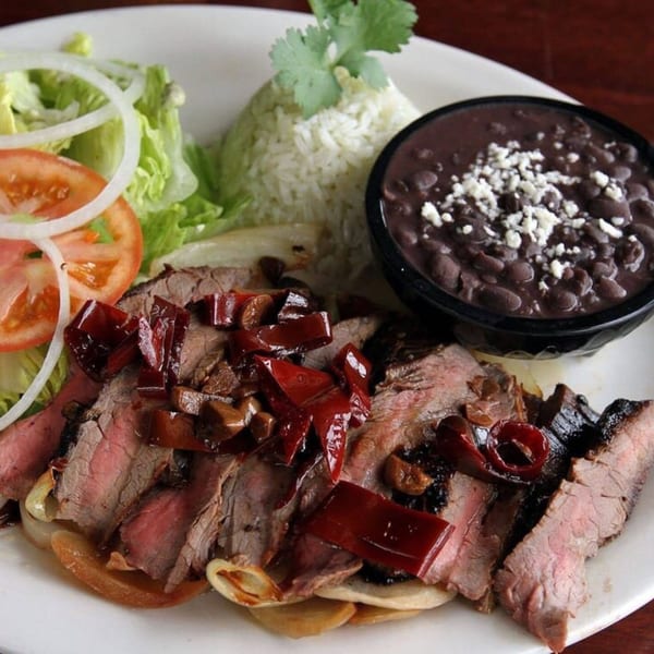Sliced steak with red peppers, onions, rice, black beans, and a side salad on a white plate.