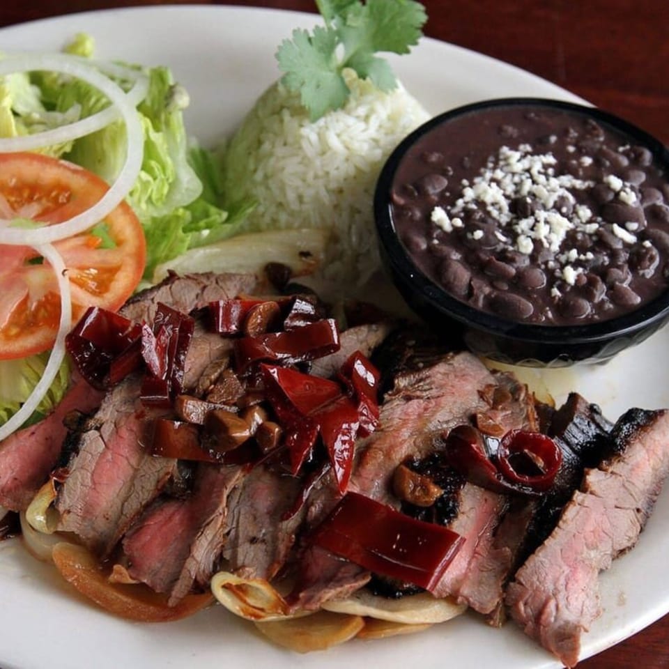 Sliced steak with red peppers, onions, rice, black beans, and a side salad on a white plate.