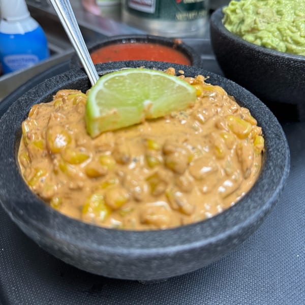 Creamy Mexican street corn dip with a lime wedge in a dark bowl, with salsa and guacamole in the background.