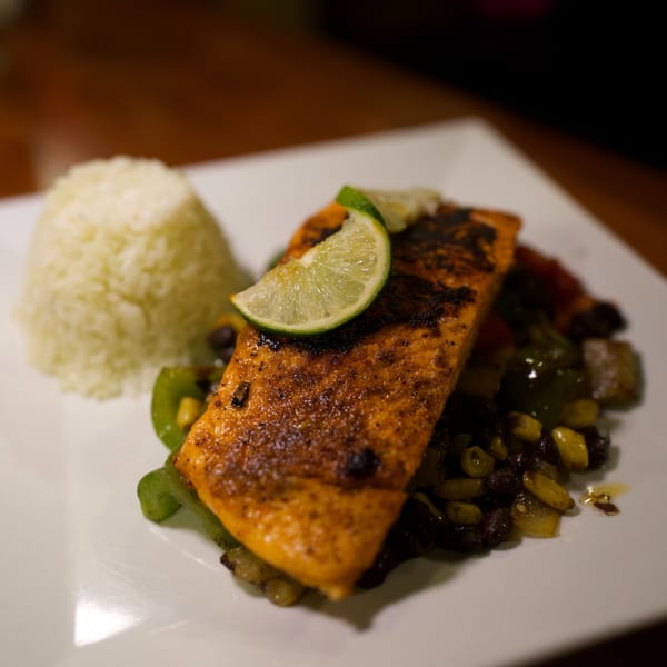 Grilled salmon with a lime wedge, black beans, corn, and bell peppers, served with white rice.