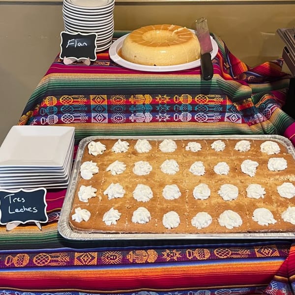 A buffet table with Flan and Tres Leches cake, adorned with a colorful Mexican blanket.