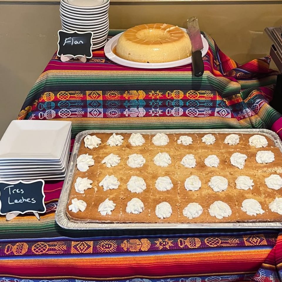 A buffet table with Flan and Tres Leches cake, adorned with a colorful Mexican blanket.
