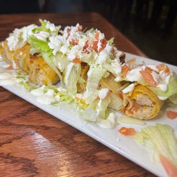 Crispy Mexican flautas topped with lettuce, diced tomatoes, sour cream, and crumbled cheese on a white plate.